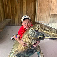 Noë participe au concours pour gagner de l'argent avec cette photo : child, boy, smiling, red_shirt, cap, bronze_statue, dinosaur, playful, indoor, wooden_walls, bench, sand_floor, sculpture, happy, posing, toy, fun, casual_clothing, cute, youth
