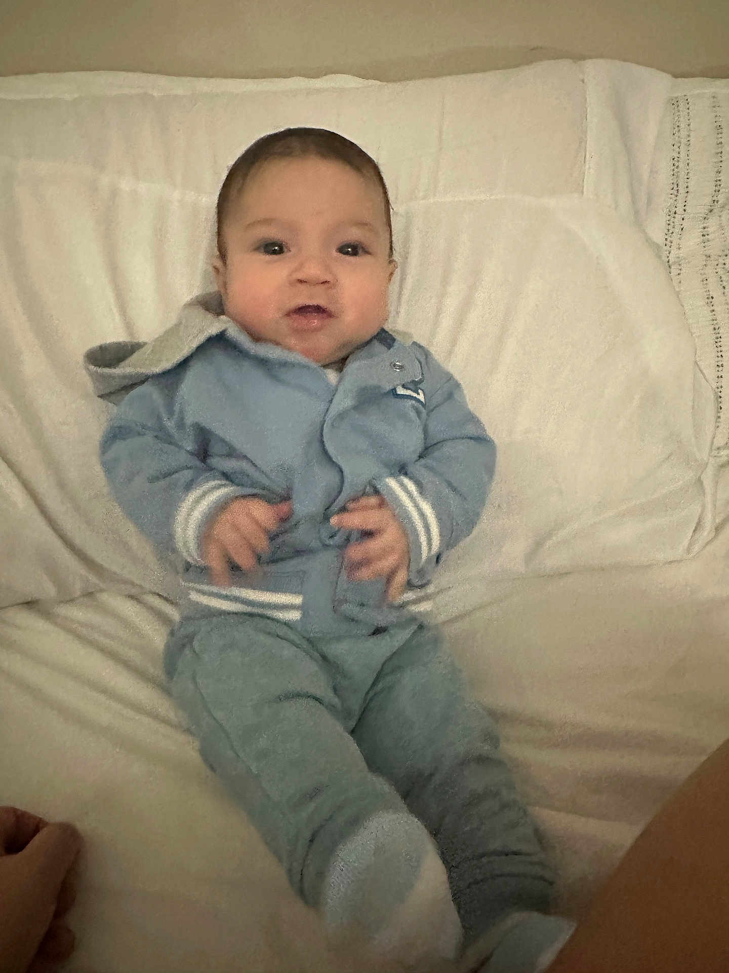 David is registered to the contest to win money with this photo: baby, infant, blue_clothing, white_blanket, sitting, hands, face, eyes, mouth, hoodie, pants, socks, indoor, person, young_child, cute, portrait, soft_lighting, cozy, casual