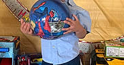 Khi is registered to the contest to win money with this photo: child, toddler, curly_hair, blue_shirt, blue_shorts, toy_ball, spiderman, toys, gift_boxes, indoor, smiling, celebration, happy, footwear, colorful, background, person, playful, party, young_child