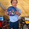 child, toddler, curly_hair, blue_shirt, blue_shorts, toy_ball, spiderman, toys, gift_boxes, indoor, smiling, celebration, happy, footwear, colorful, background, person, playful, party, young_child