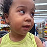toddler, child, face, curly_hair, green_tank_top, indoor, store, aisle, shelves, products, shopping, casual_clothing, portrait, young_child, cute, curious, person, people, background_blur, daylight
