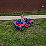 child, toddler, toy_car, ride_on_toy, red_car, blue_car, grass, lawn, brick_wall, sidewalk, window, smiling, portrait, outdoor, playing, wheels, steering_wheel, happy, casual_clothing, suburban