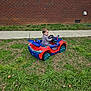 child, toddler, toy_car, grass, sidewalk, brick_wall, window, outdoor, playing, smiling, steering_wheel, blue, red, wheels, lawn, concrete, person, vehicle, portrait, casual_clothing