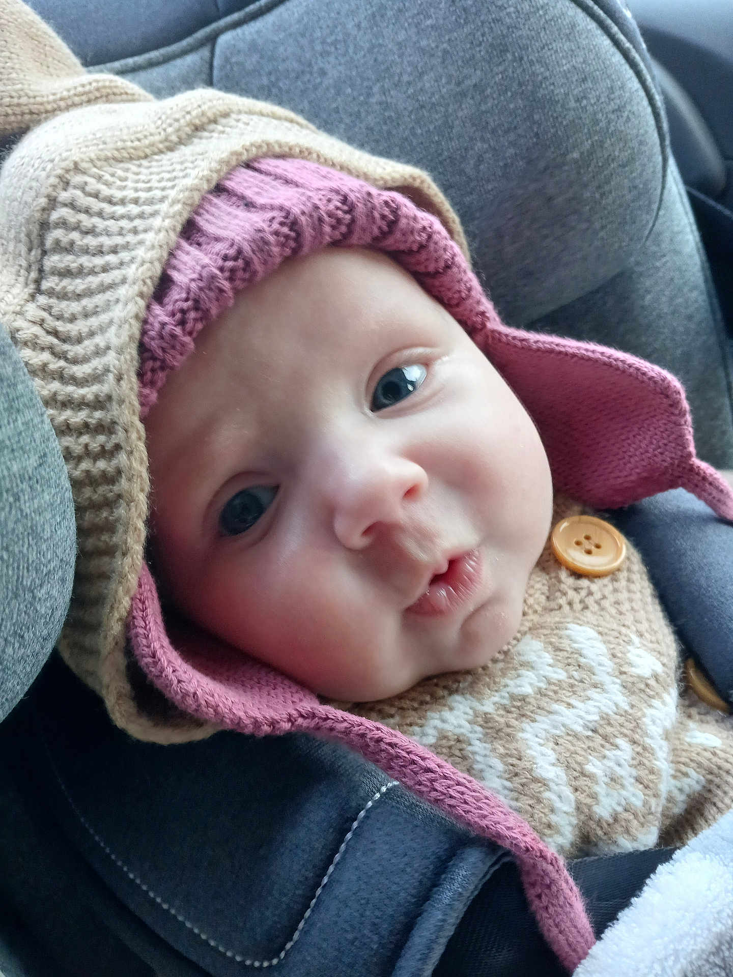 Lynaë a rejoint le concours — aidez-le/la à gagner de superbes lots ! baby, face, closeup, knitted_hat, knitted_sweater, button, seatbelt, car_seat, child, cute, expression, warm_clothing, indoors, infant, portrait, person, headwear, seat, young_child, soft_lighting