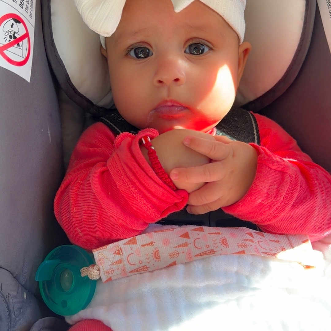 Aurora is registered to the contest to win money with this photo: baby, infant, child, car_seat, bow, headband, red_clothing, white_blanket, pacifier, portrait, face, eyes, sitting, sunlight, indoors, travel, vehicle, hands, baby_clothing, photography