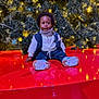 child, christmas_tree, cute, decorations, happy, holiday_lights, indoor, jeans, ornament, pine_branches, portrait, red_platform, reflection, scarf, seasonal, sitting, smile, sneakers, toddler, vest