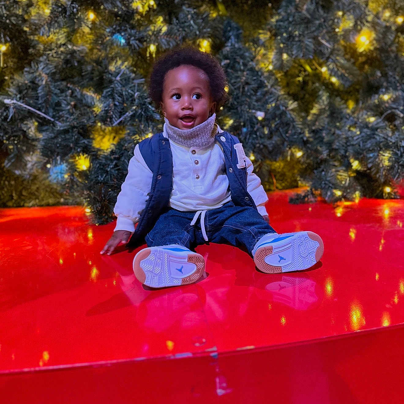 Jaylon a rejoint le concours — aidez-le/la à gagner de superbes lots ! child, christmas_tree, cute, decorations, happy, holiday_lights, indoor, jeans, ornament, pine_branches, portrait, red_platform, reflection, scarf, seasonal, sitting, smile, sneakers, toddler, vest