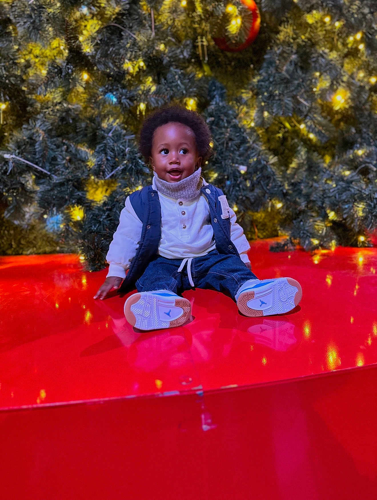 Jaylon a rejoint le concours — aidez-le/la à gagner de superbes lots ! child, christmas_tree, cute, decorations, happy, holiday_lights, indoor, jeans, ornament, pine_branches, portrait, red_platform, reflection, scarf, seasonal, sitting, smile, sneakers, toddler, vest