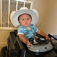 Gabe is registered to the contest to win money with this photo: child, toddler, cowboy_hat, toy_car, indoor, steering_wheel, gear_shift, blue_shirt, short_sleeves, serious_expression, face, hand, seat, vent, wall, floor, toy, vehicle, person, baby