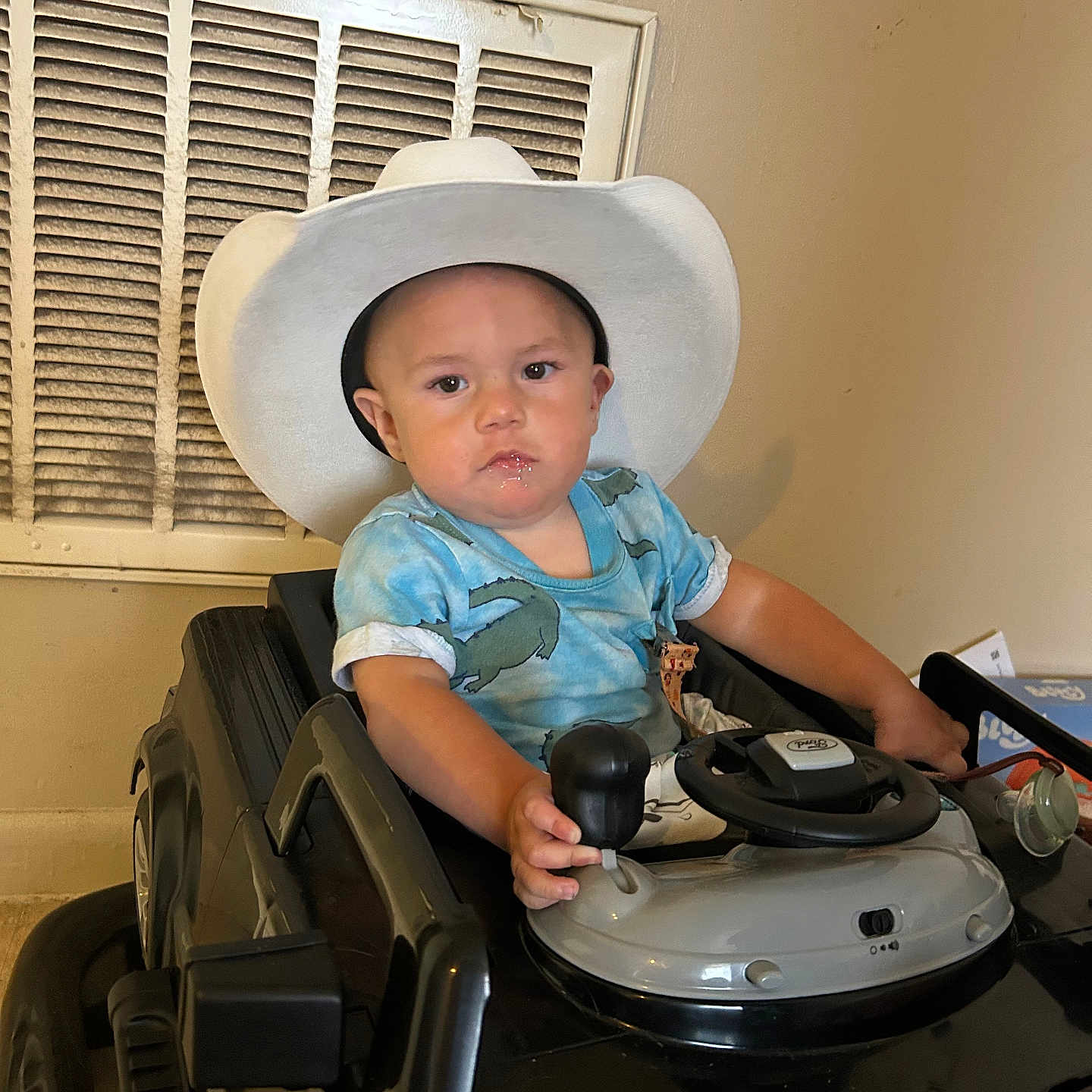 Gabe is registered to the contest to win money with this photo: baby, blue_shirt, child, cowboy_hat, face, floor, gear_shift, hand, indoor, person, seat, serious_expression, short_sleeves, steering_wheel, toddler, toy, toy_car, vehicle, vent, wall