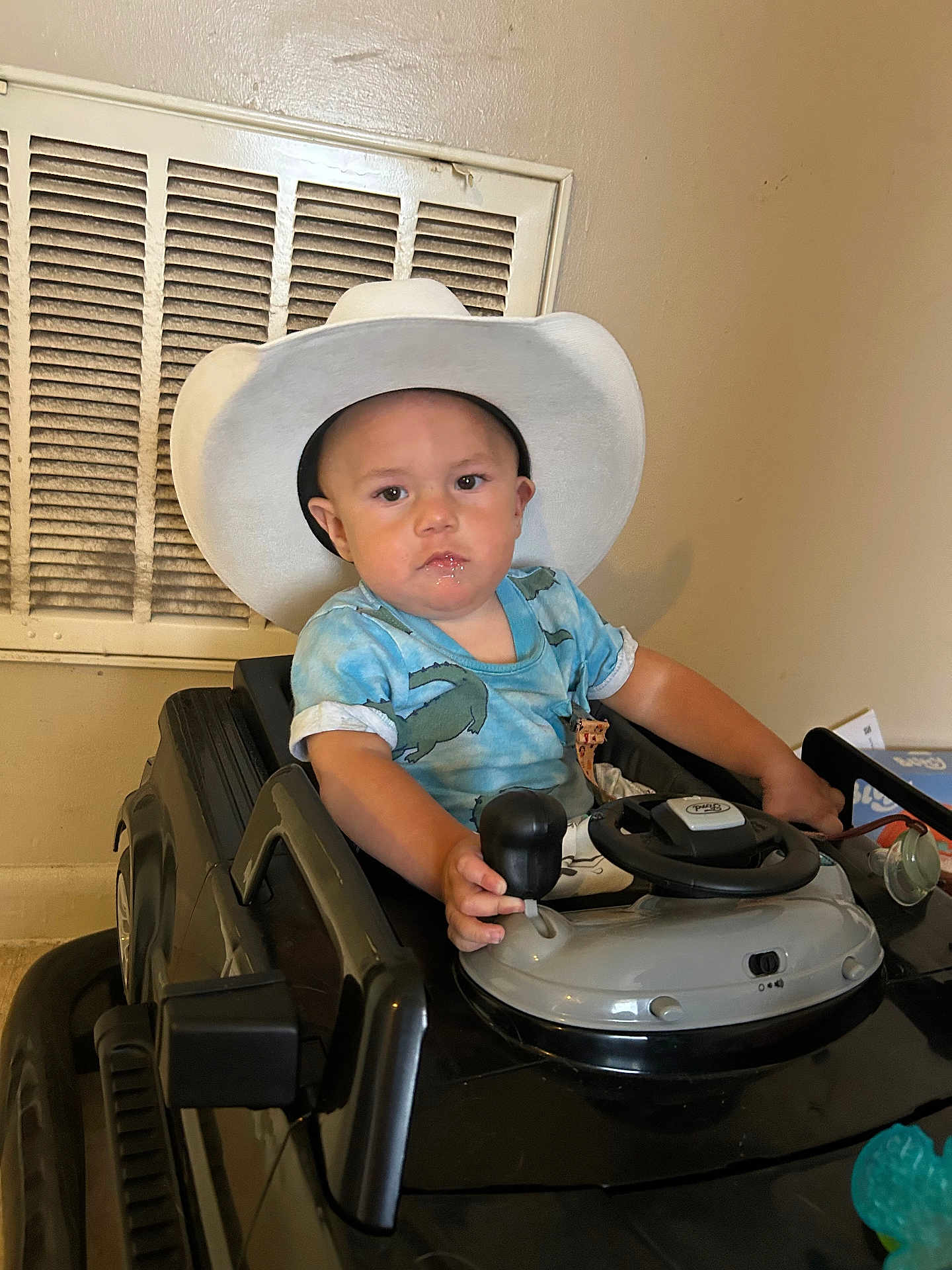 Gabe is registered to the contest to win money with this photo: child, toddler, cowboy_hat, toy_car, indoor, steering_wheel, gear_shift, blue_shirt, short_sleeves, serious_expression, face, hand, seat, vent, wall, floor, toy, vehicle, person, baby