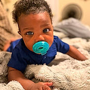 Jiovonni is registered to the contest to win money with this photo: baby, child, infant, pacifier, curly_hair, blue_shirt, portrait, close_up, smiling, face, eyes, bedroom, bed, blanket, bedding, cozy, indoor, sitting, crawling, photography