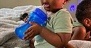 Jiovonni is registered to the contest to win money with this photo: child, toddler, baby, siblings, bottle, drinking, bed, blanket, bedroom, portrait, sitting, curly_hair, striped_shirt, green_clothing, blue_bottle, cozy, indoors, family, playful, close_up