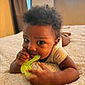 baby, child, infant, portrait, indoors, bed, bedspread, lying_down, teething_toy, green_toy, chewing, curly_hair, brown_eyes, curious, cozy, home, television, warm_lighting, close_up, smiling