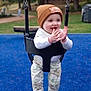 baby, child, swing, playground, blue_floor, beanie, clapping_hands, happy, outdoor, trees, park, infant, clothing, camo_pants, socks, smiling, seat, fun, daytime, nature