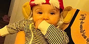 Cairo is registered to the contest to win money with this photo: baby, child, elf_hat, striped_clothing, hand_sucking, adult_arm, tattoo, indoor, person, cute, costume, red_hat, white_fluff, beige_ears, holding, portrait, close_up, warm_lighting, human, casual