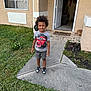 child, boy, smiling, standing, tshirt, shorts, sneakers, grass, concrete_path, house, door, window, cat, sidewalk, outdoor, daytime, curly_hair, happy, residential, front_yard