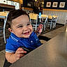 baby, child, smiling, blue_shirt, high_chair, restaurant, table, indoor, furniture, happy, face, person, cute, dining, seat, portrait, food, hands, expression, casual