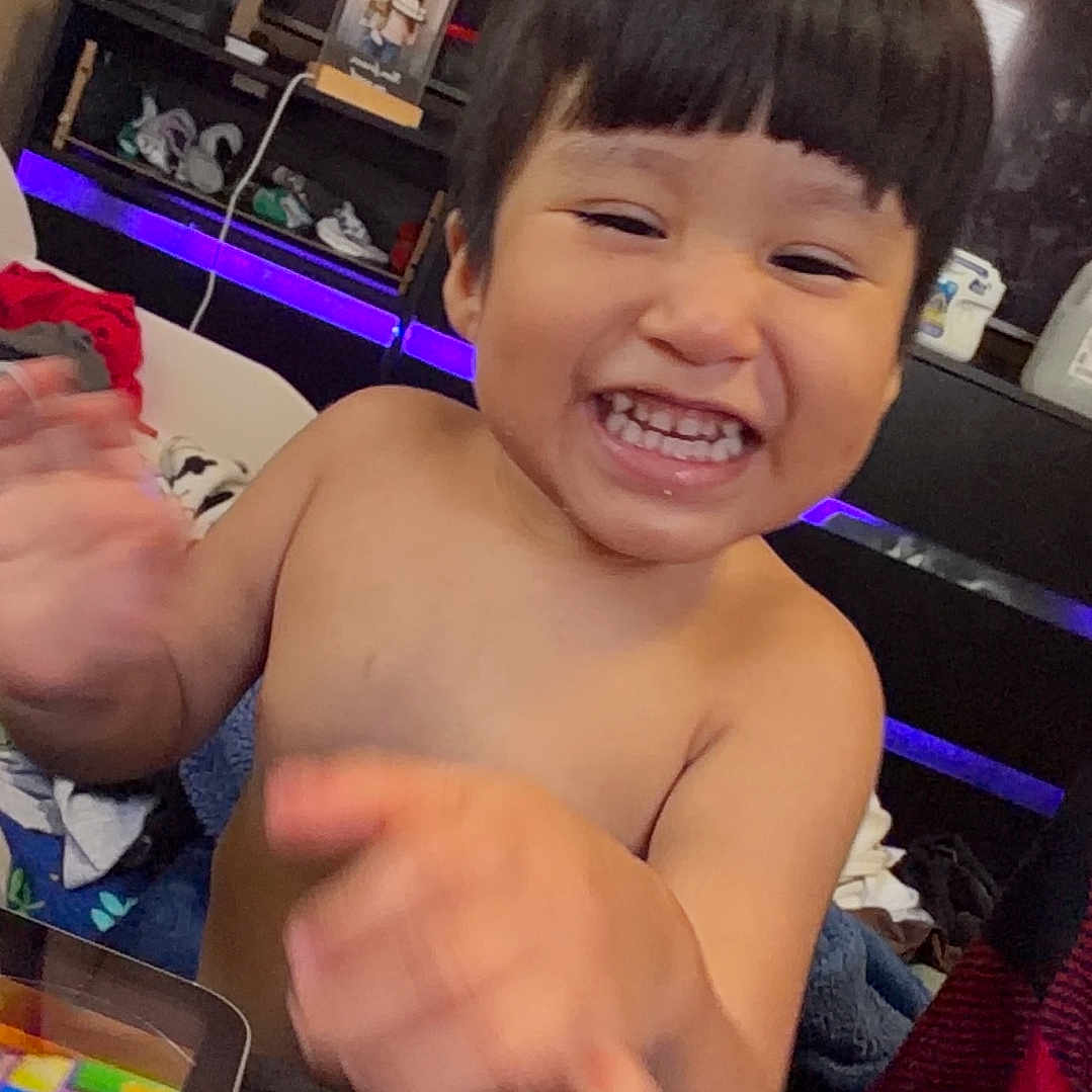 Liam Rivera is registered to the contest to win money with this photo: child, toddler, smiling, laughing, happy, portrait, indoors, home, sitting, waving, hands, short_hair, bangs, bare_shoulders, clothing, toy, tablet, television, led_lights, furniture