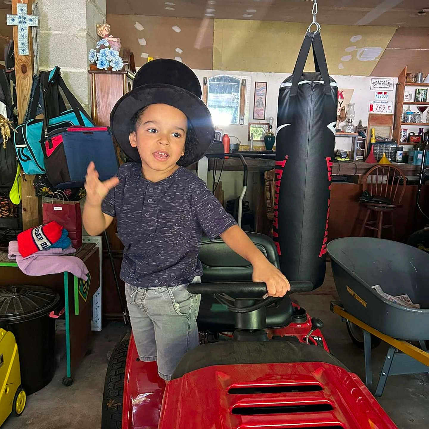 Michael joined the competition — help win amazing prizes! child, young_child, hat, black_hat, smiling, gesturing, portrait, outdoor_indoor, garage, workshop, riding_mower, red_vehicle, punching_bag, furniture, chairs, tools, storage, cross, clothing, photography