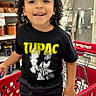 child, toddler, smiling, curly_hair, portrait, standing, shopping_cart, grocery_store, supermarket, retail, indoor, shelves, groceries, red_cart, black_clothing, graphic_t_shirt, tupac_shirt, store_lighting, happy, shopping