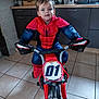 child, toddler, boy, spiderman_costume, costume, toy_bike, tricycle, indoor, kitchen, cabinet, countertop, tiled_floor, portrait, face, smiling, brown_hair, eyes, red, blue, number_plate