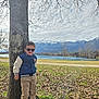 child, boy, sunglasses, tree, tree_trunk, jacket, cargo_pants, boots, park, lake, mountains, snow_capped_mountains, sky, clouds, grass, fallen_leaves, smile, portrait, outdoors, sunlight