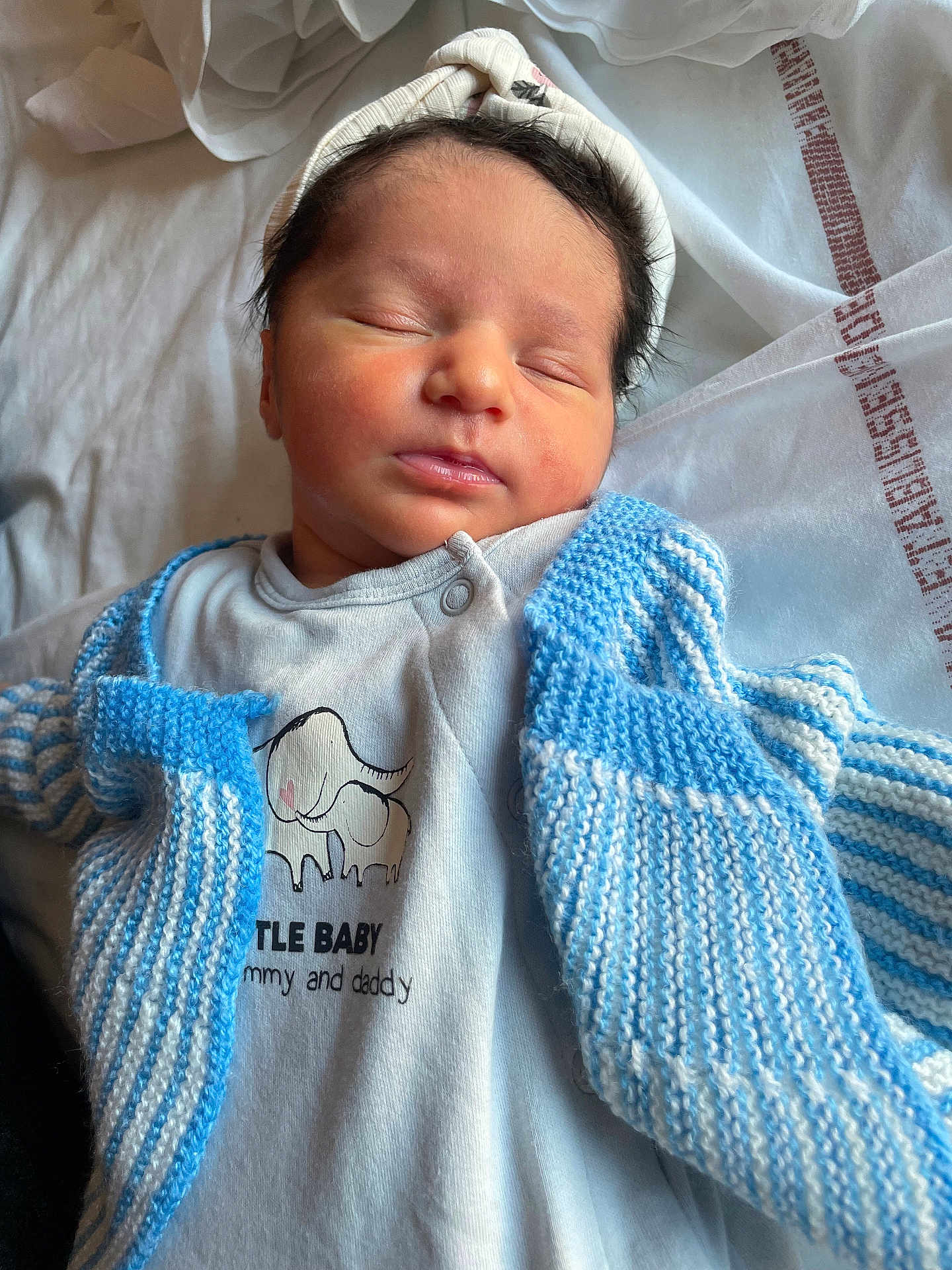 Lùcia a rejoint le concours — aidez-le/la à gagner de superbes lots ! newborn, baby, sleeping, infant, knitted_cardigan, blue, white, onesie, elephant_graphic, text_on_clothing, soft_blanket, peaceful, cute, portrait, closeup, headband, clothing, indoors, child, resting