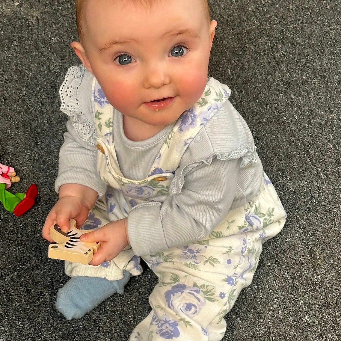 Coralie joined the competition — help win amazing prizes! baby, blue_socks, carpet, child, clothing, curious, cute, face, floor, floral, hands, indoor, infant, overalls, person, playthings, sitting, smiling, toy, wooden_toy