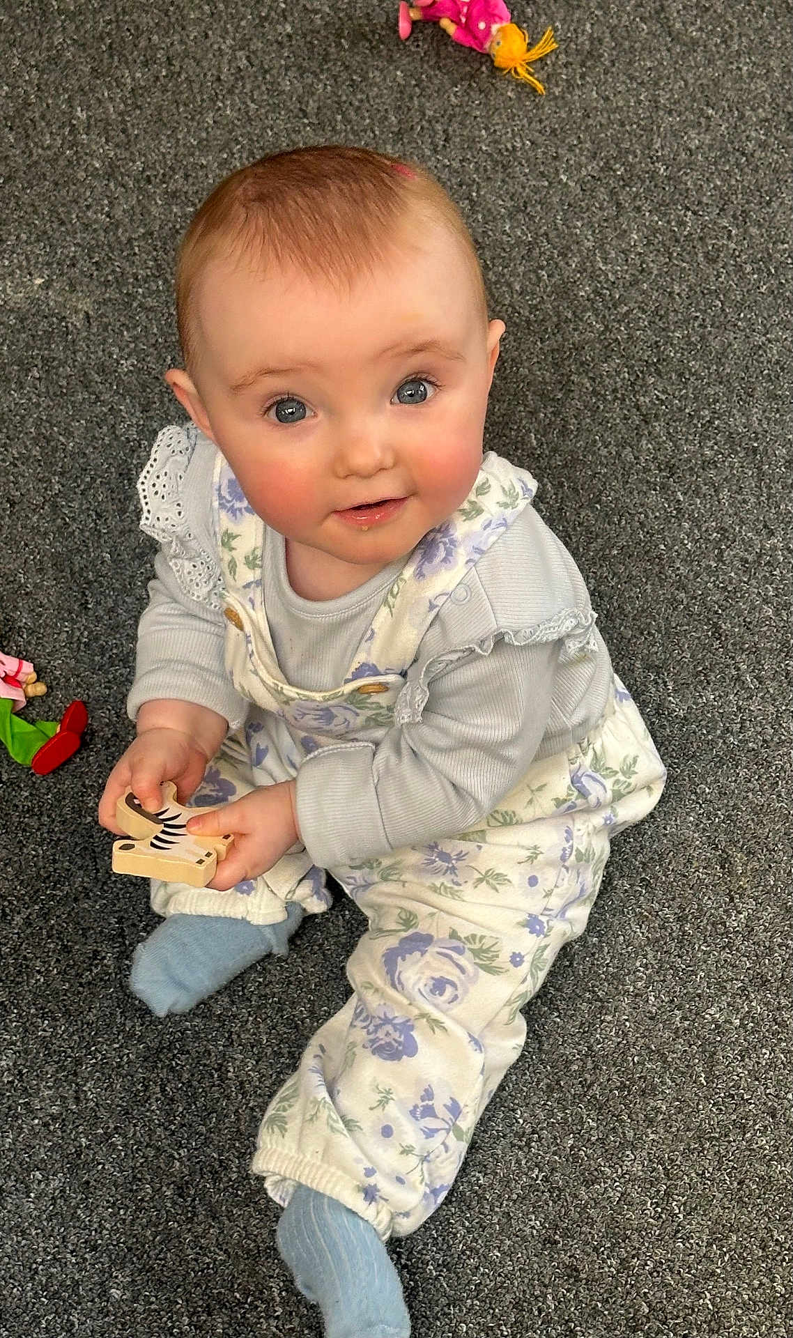 Coralie joined the competition — help win amazing prizes! baby, child, carpet, toy, wooden_toy, blue_socks, overalls, floral, playthings, indoor, cute, smiling, curious, sitting, infant, face, hands, clothing, person, floor