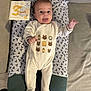 baby, infant, onesie, cushion, milestone_card, tongue_out, patterned_fabric, smiling, lying_down, cute, child, person, home, soft_lighting, decorative_clothing, plush_surface, happy, face, small_hands, footed_pajamas
