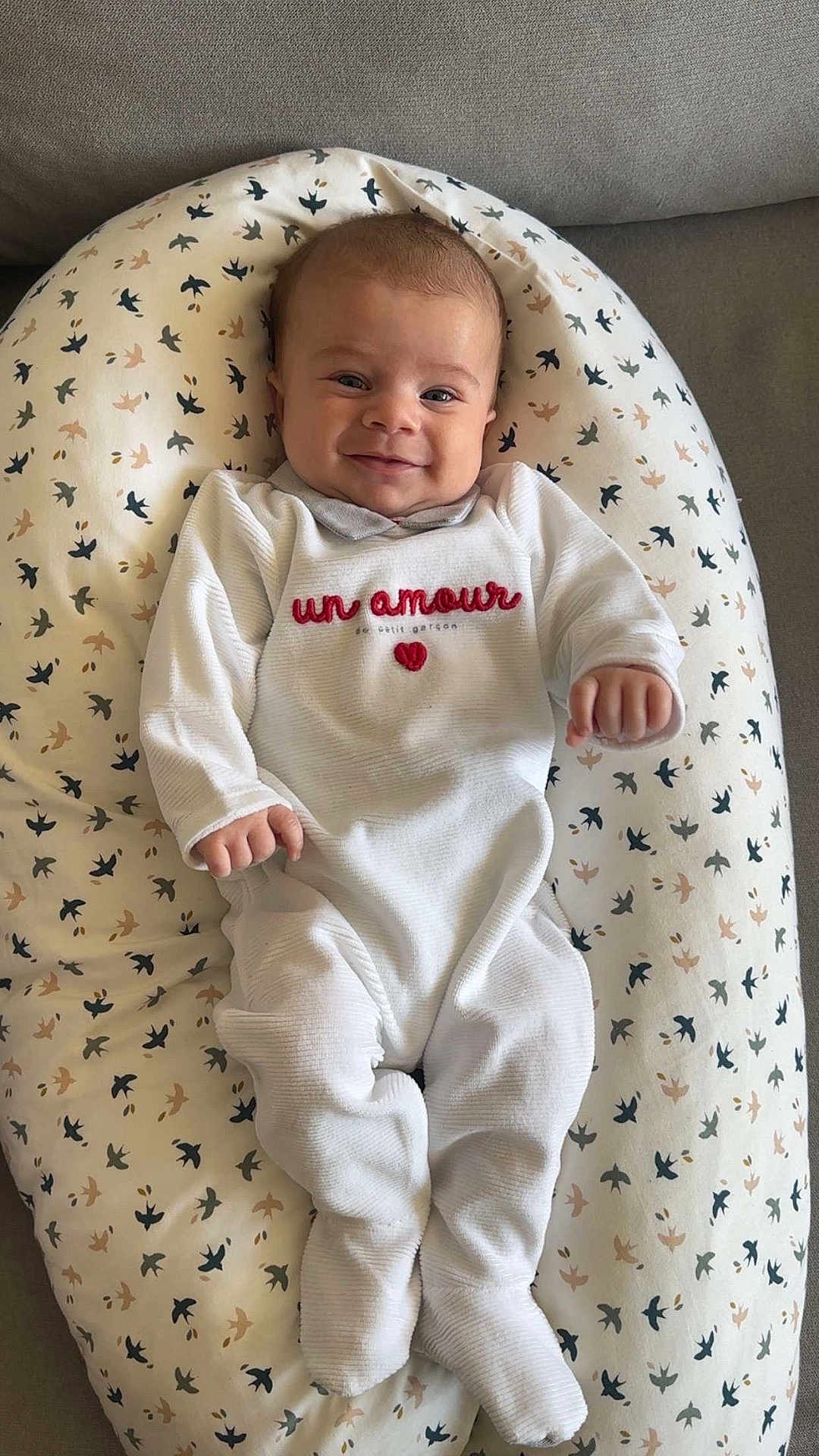 Gabriel participe au concours pour gagner de l'argent avec cette photo : baby, infant, smiling, onesie, white_clothing, cushion, bird_pattern, soft_furniture, indoor, child, portrait, cute, happy, lying_down, cozy, small_hands, head, face, blue_eyes, toddler