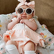 Amelia is registered to the contest to win money with this photo: baby, infant, sunglasses, headband, bow, blanket, floral_pattern, sitting, cute, fashion, clothing, white_socks, soft_lighting, portrait, indoor, couch, smile, hands, legs, skin