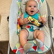 Giovanni is registered to the contest to win money with this photo: baby, infant, baby_seat, popcorn, toy, colorful, patterned, happy, smiling, indoor, floor, carpet, clothing, socks, face, child, cute, playful, seated, holding