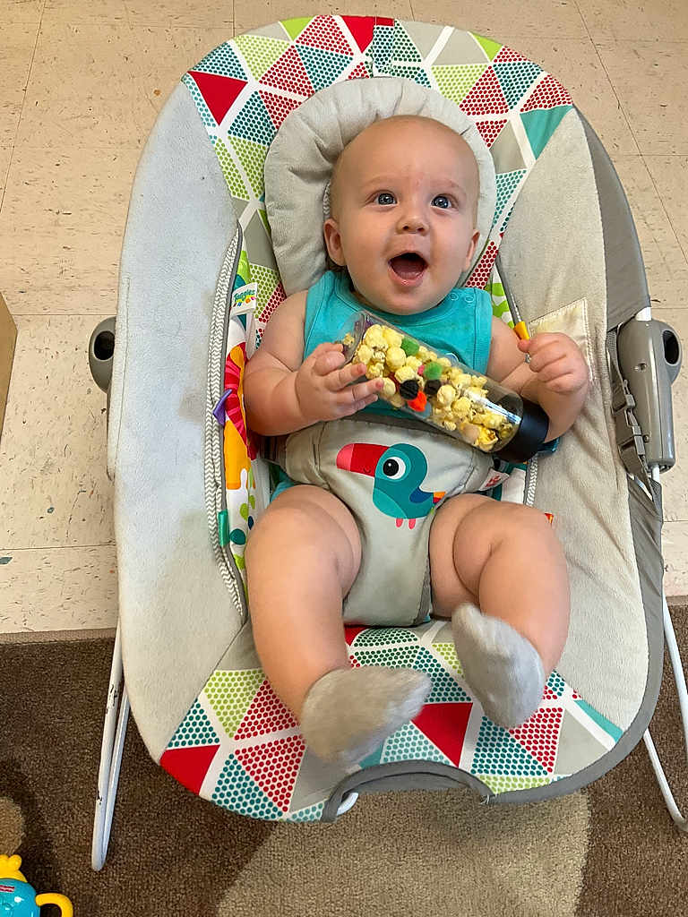 Giovanni is registered to the contest to win money with this photo: baby, infant, baby_seat, popcorn, toy, colorful, patterned, happy, smiling, indoor, floor, carpet, clothing, socks, face, child, cute, playful, seated, holding