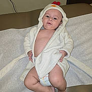 Declan is registered to the contest to win money with this photo: baby, child, clothing, cute, diaper, expression, feet, hand, hood, indoors, infant, laying, medical_table, person, portrait, relaxing, robe, skin, toddler, young