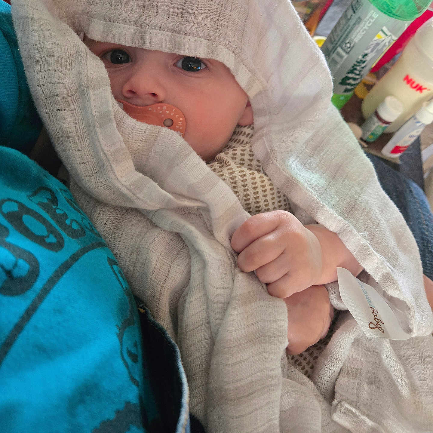 Declan is registered to the contest to win money with this photo: baby, blanket, pacifier, hand, face, person, clothing, fabric, indoor, bottle, can, table, container, green_bottle, blue_clothing, soft_texture, infant, holding, wrapped, closeup