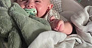 Declan is registered to the contest to win money with this photo: baby, pacifier, blanket, plush_toy, smiling, cozy, infant, face, hand, fabric, comfort, cute, bed, sleeping_area, soft, person, young_child, indoors, happy, resting