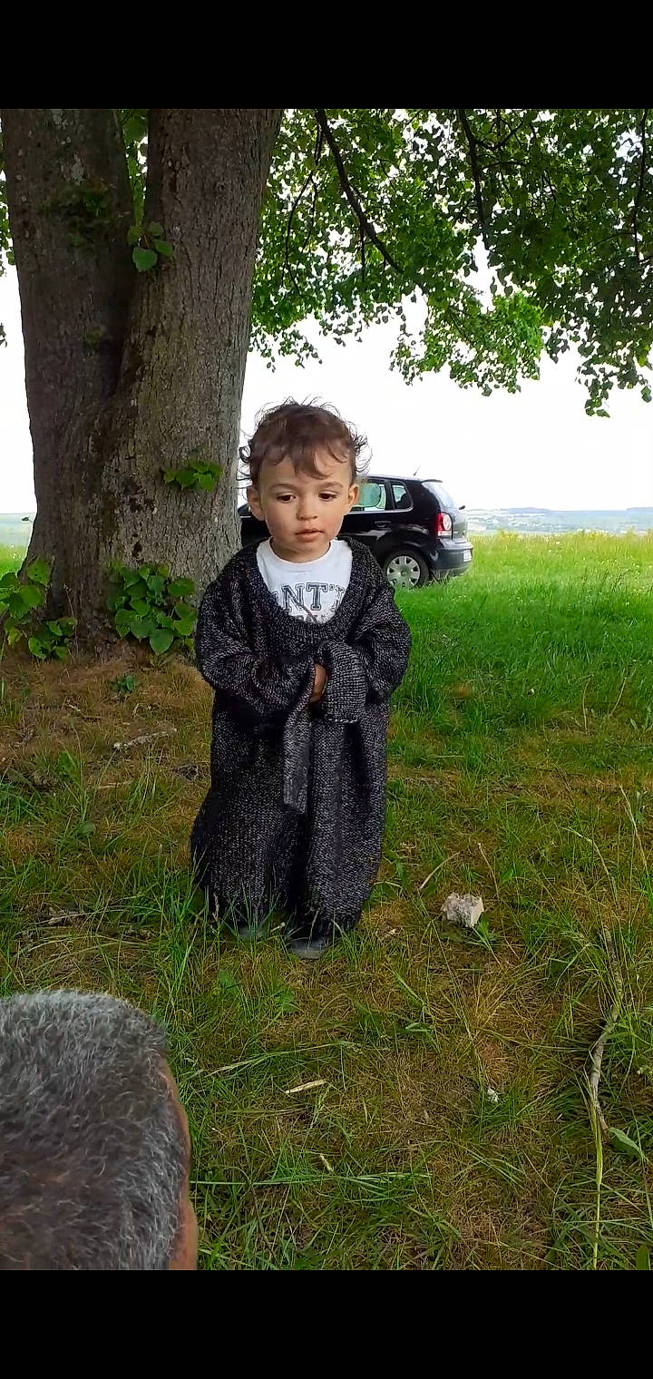 Ismaël participe au concours pour gagner de l'argent avec cette photo : car, denim, fur, grass, grassland, jacket, lawn, pattern, people_in_nature, person, plaid, plant, prairie, sitting, sky, soil, tire, toddler, tree, trunk
