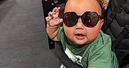 Ethan is registered to the contest to win money with this photo: baby, child, sunglasses, smiling, thumbs_up, stroller, restaurant, indoor, red_chairs, table, green_shirt, seated, portrait, happy, infant, casual, fashion, playful, dining_area, photograph