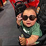 baby, child, sunglasses, smiling, thumbs_up, stroller, restaurant, indoor, red_chairs, table, green_shirt, seated, portrait, happy, infant, casual, fashion, playful, dining_area, photograph