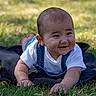 baby, smiling, grass, outdoor, child, infant, denim_overalls, white_shirt, happy, cute, face, earrings, skin, lying_down, nature, sunlight, greenery, toddler, black_cloth, portrait