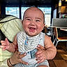 baby, smiling, bib, infant, person, indoor, restaurant, holding, happy, cute, child, clothing, table, chair, floor, window, light, skin, hand, adult