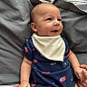 Colter is registered to the contest to win money with this photo: baby, infant, smiling, onesie, bib, hand_holding, bed, bedding, gray_sheet, striped_sheet, person, cute, portrait, child, indoor, happy, skin, face, small_hands, comfort