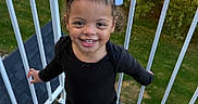 Nova Sage is registered to the contest to win money with this photo: child, toddler, girl, smiling, portrait, outdoor, deck, balcony, railing, bows, hair_accessories, curly_hair, black_clothing, white_shoes, standing, happy, teeth, garden, greenery, daylight