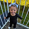 child, toddler, girl, smiling, portrait, outdoor, deck, balcony, railing, bows, hair_accessories, curly_hair, black_clothing, white_shoes, standing, happy, teeth, garden, greenery, daylight
