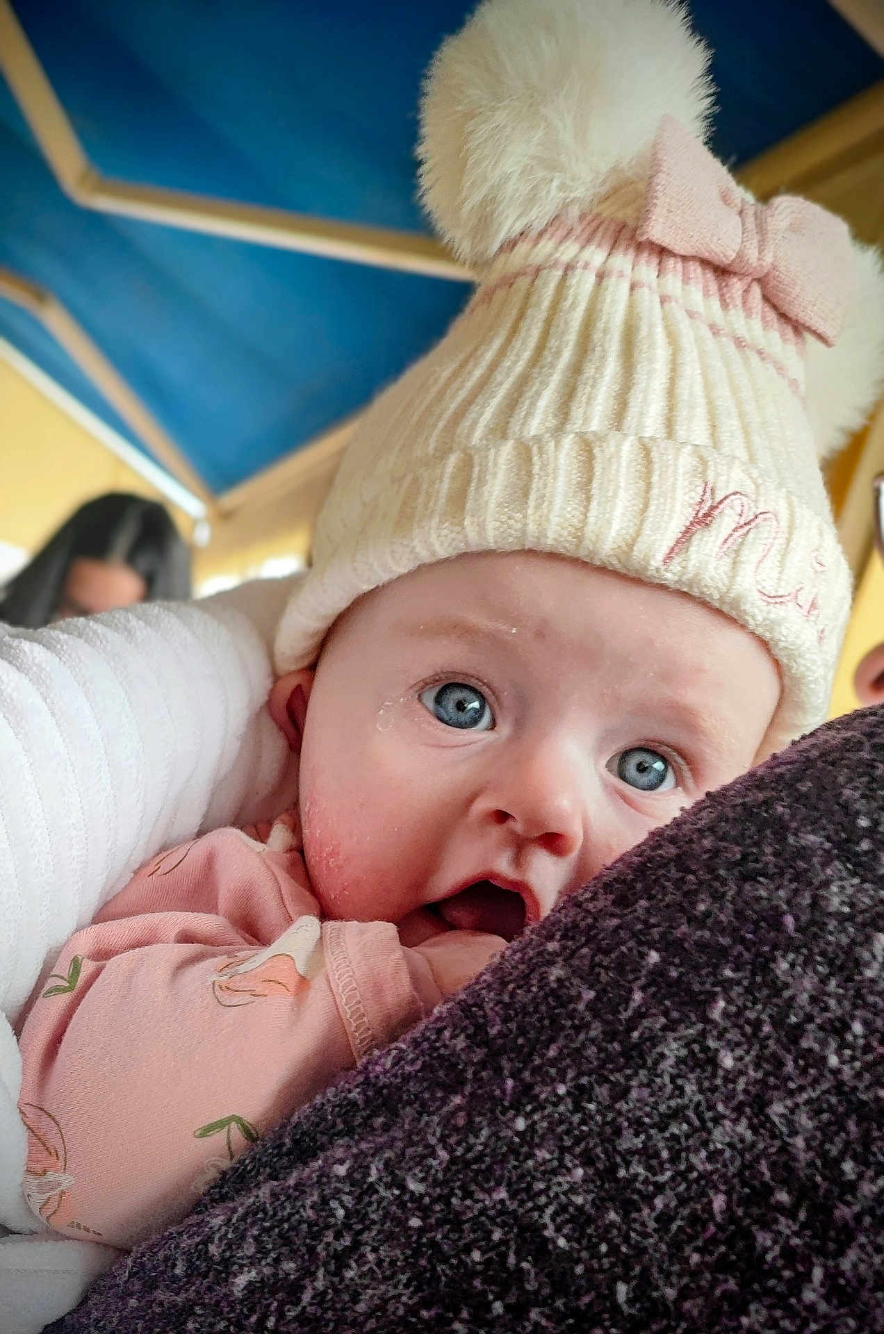 Louise participe au concours pour gagner de l'argent avec cette photo : baby, infant, child, blue_eyes, knit_hat, pom_pom, bow, pink_onesie, close_up, portrait, surprised_expression, cheeks, drool, skin_detail, blanket, arm, adult_in_background, canopy, indoor, soft_texture