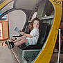 child, smiling, curly_hair, helicopter, yellow, cockpit, seat, indoor, booklet, person, play_area, museum, exhibit, metal, footwear, black_shoes, white_shirt, happy, curious, background