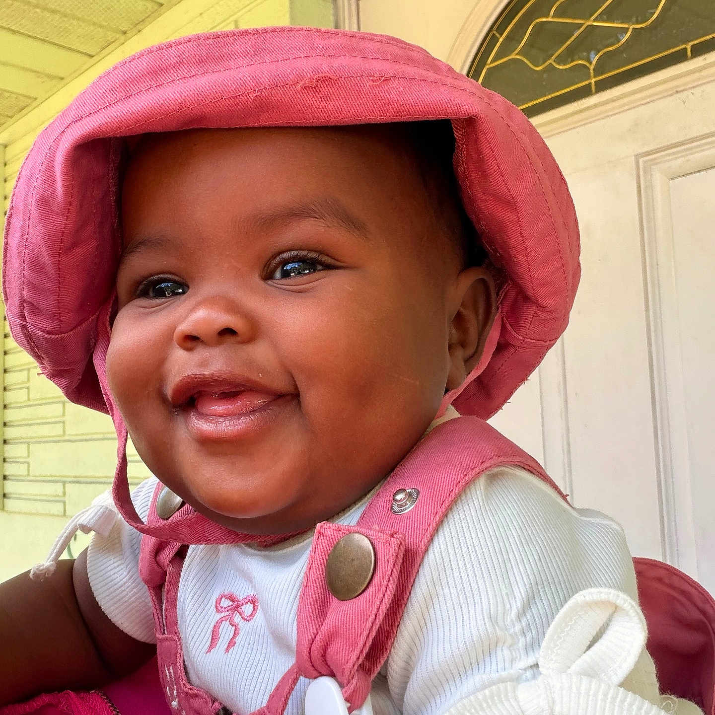 Kulture is registered to the contest to win money with this photo: baby, child, smiling, pink_hat, pink_overalls, white_shirt, happy, face, portrait, indoors, door, decorative_glass, closeup, cute, infant, cheerful, person, clothing, babywear, headwear