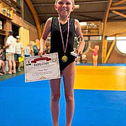 Nancy participe au concours pour gagner de l'argent avec cette photo : child, girl, gymnast, leotard, medal, trophy, diploma, award, winning, smile, barefoot, blue_mat, yellow_mat, indoor_gym, spectators, wooden_beams, pose, celebration, portrait, blurred_background